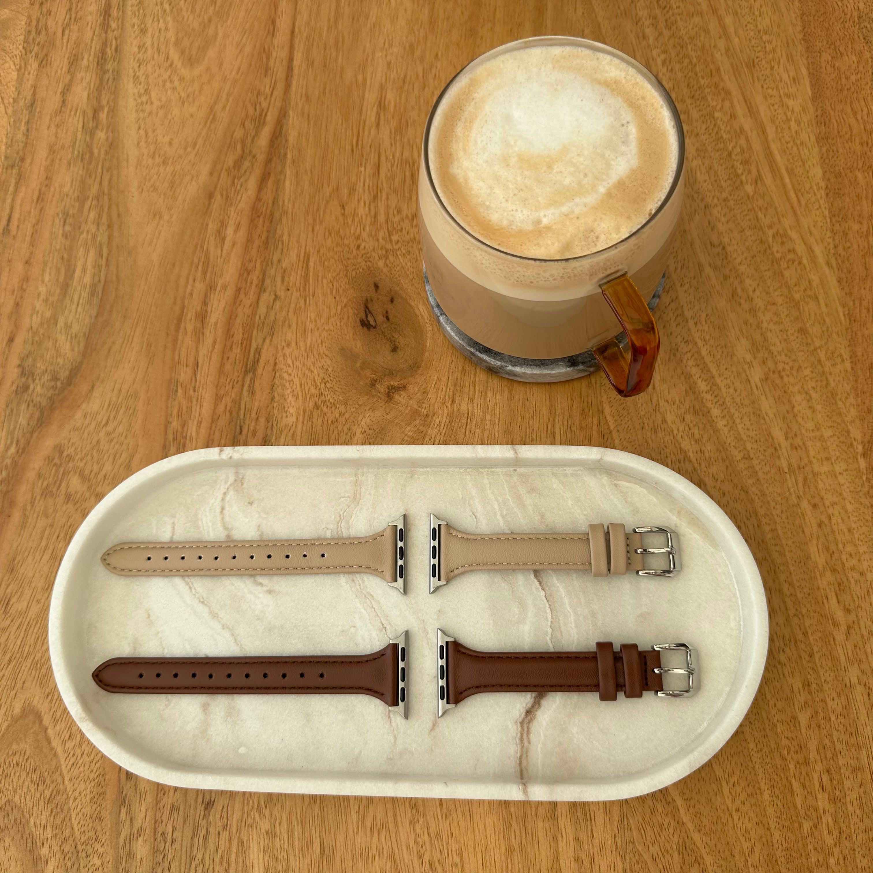 Two Lisse Apple watch straps on a marble tray with a glass of cappuccino on a wooden table.